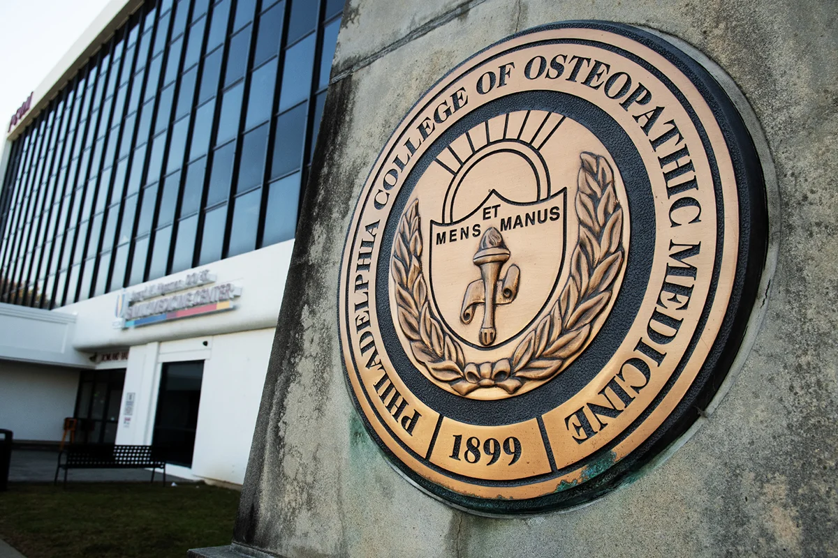 PCOM obelisk and College seal in the foreground with the Hassman Family Medicine Center entranceway in the background
